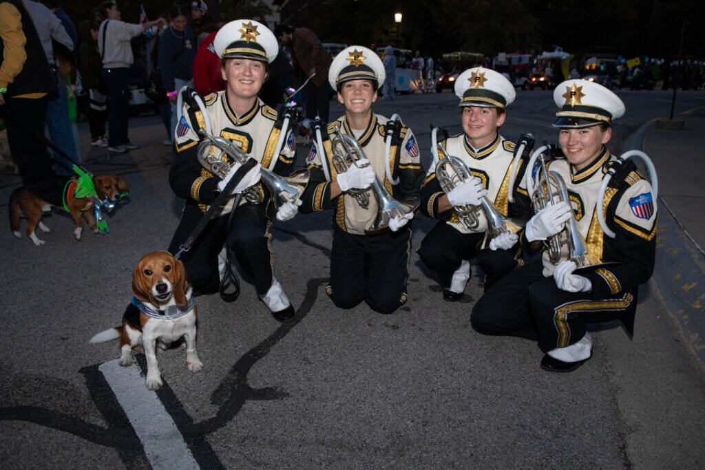 Four Purdue University band members, dressed in full black and gold uniform, are posing for a photo with a beagle, who is sitting on the far left side of the photo.