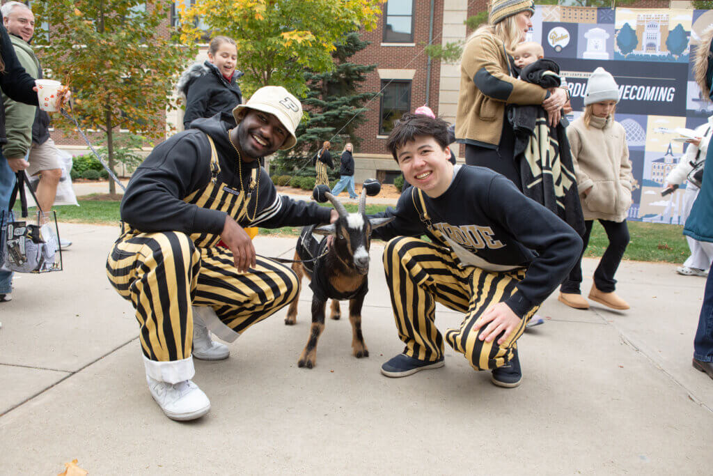 Two men wearing gold and black overalls are kneeling down to pose for a photo with a goat