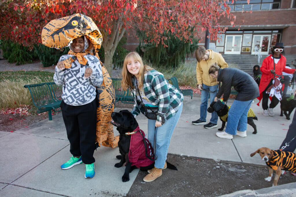 A girl in a blue and white plaid shirt is posing outdoors with a black-colored dog, and a girl in a dinosaur costume is smiling at the camera. There are other dogs and people in the background.