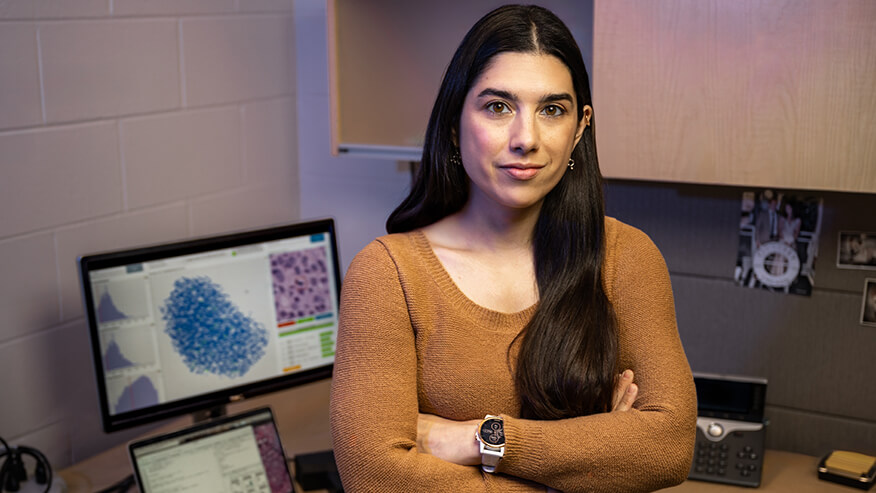 A person with long dark hair stands with folded arms in an office, with computer screens displaying scientific data behind them.