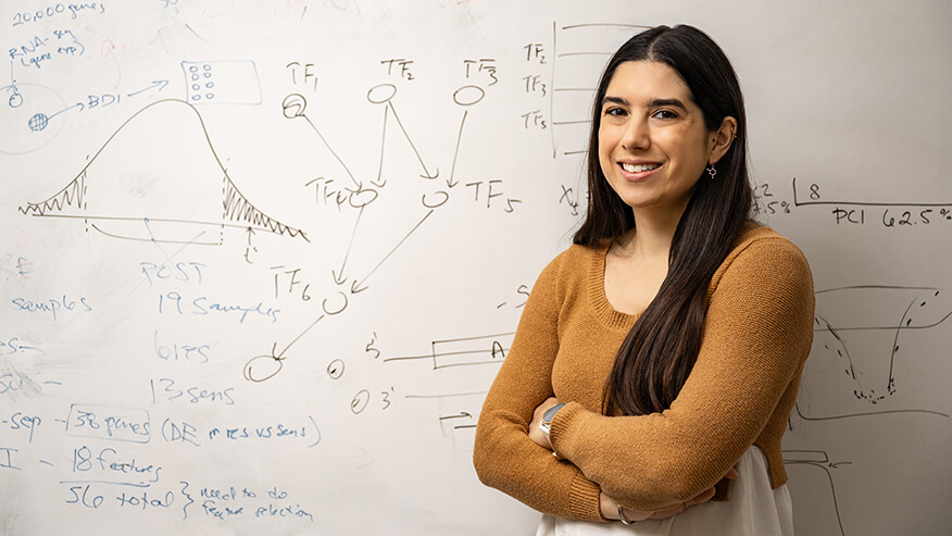 A person with long dark hair wearing a yellow-brown sweater, standing with their arms crossed in front of a white board with scientific notes written on it.