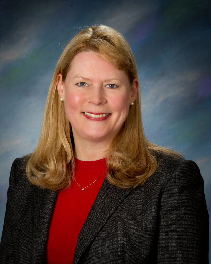 portrait of a woman facing the camera, smiling. The woman is wearing a red shirt with a black jacket and has long hair. The photo has a dark blue background