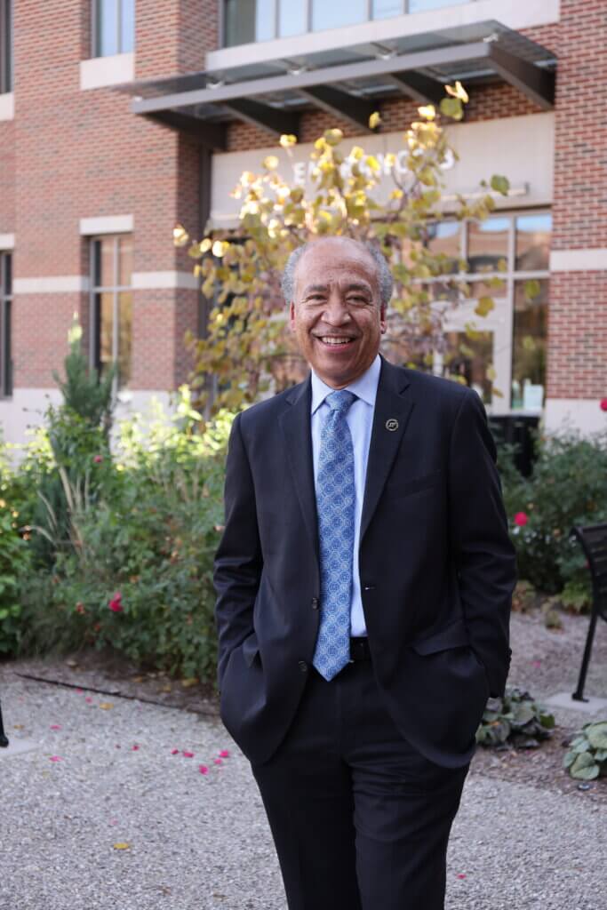 portrait of man standing outdoors, in front of a flower garden and brick building