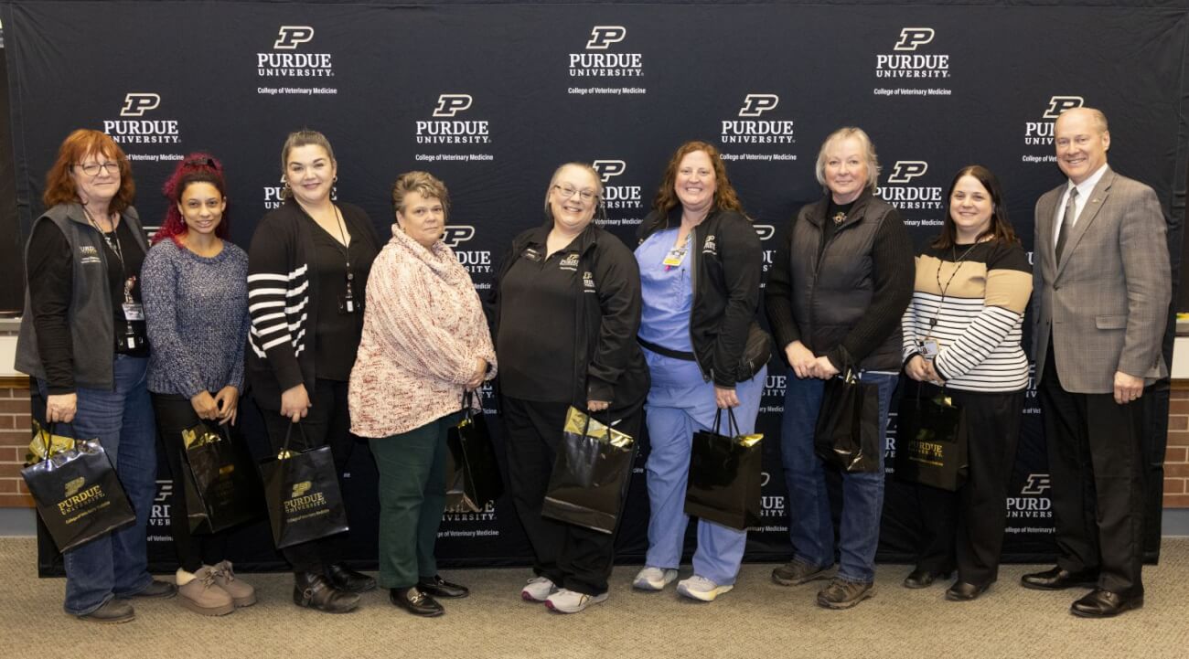 A group of nine people standing for a photo in front of a large black Purdue University background