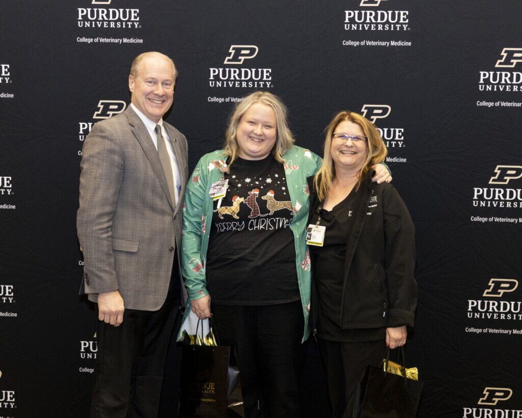 Three people stand in front of a black Purdue University backdrop for a photo. The person on the far left is a man wearing a gray suit jacket, the woman in the middle is wearing Christmas-themed attire, and the woman on the right is wearing glasses and a black shirt.