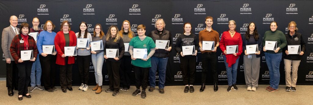 A large group of people, both men and women, stand holding their awards in front of a large black Purdue University backdrop