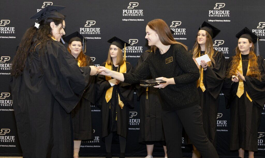 Two women are exchanging an item in front of a group of graduating students. Everyone in the photo is wearing black and the students in the background have gold sashes
