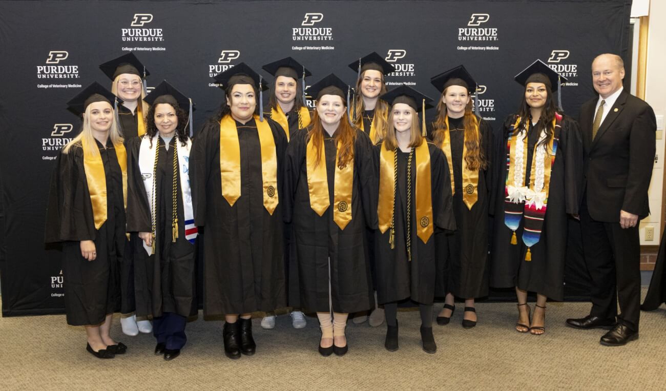 A group of graduating students stand in front of a large black backdrop. They are wearing black gowns with gold sashes