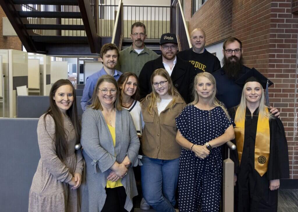 A group of proud-looking family members stand with a graduating student, who is on the far right of the photo in a black gown and gold sash. There is a staircase behind the group.
