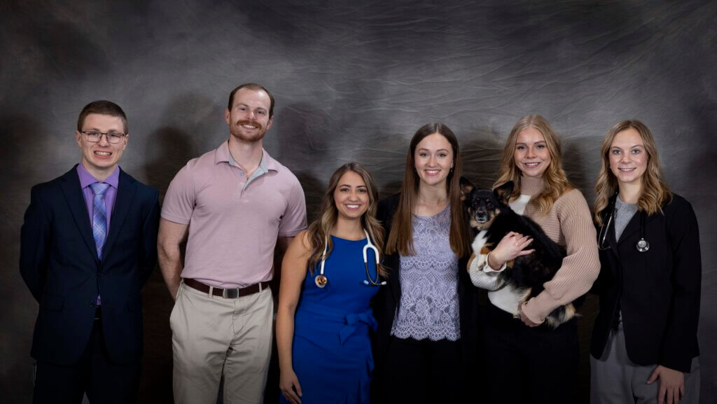 A group of six young people stand in a line in front of a gray backdrop. They are wearing a variety of colors and are all dressed in business casual attire. One of the women on the right side of the photo is holding a small black dog.