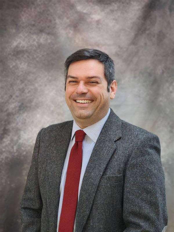 A portrait of a man who is smiling and wearing a gray suit jacket and a red tie. The photo includes a gray background.