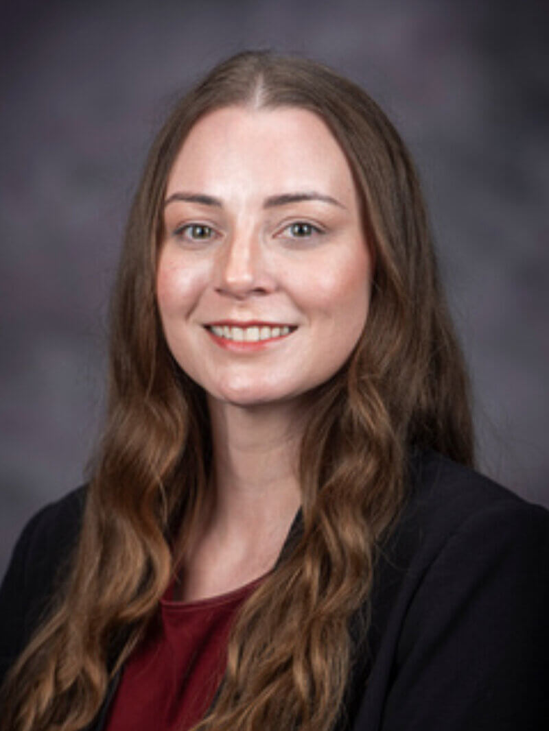 portrait of a woman with long brown hair, wearing a red shirt and a black jacket and is in front of a gray background