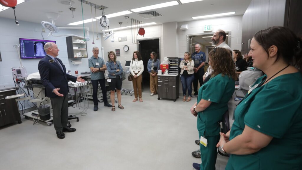 A large group of people are standing on the right side of the photo, observing a man positioned on the left side of the photo speak to them. The man is wearing a black suit with his arms out.