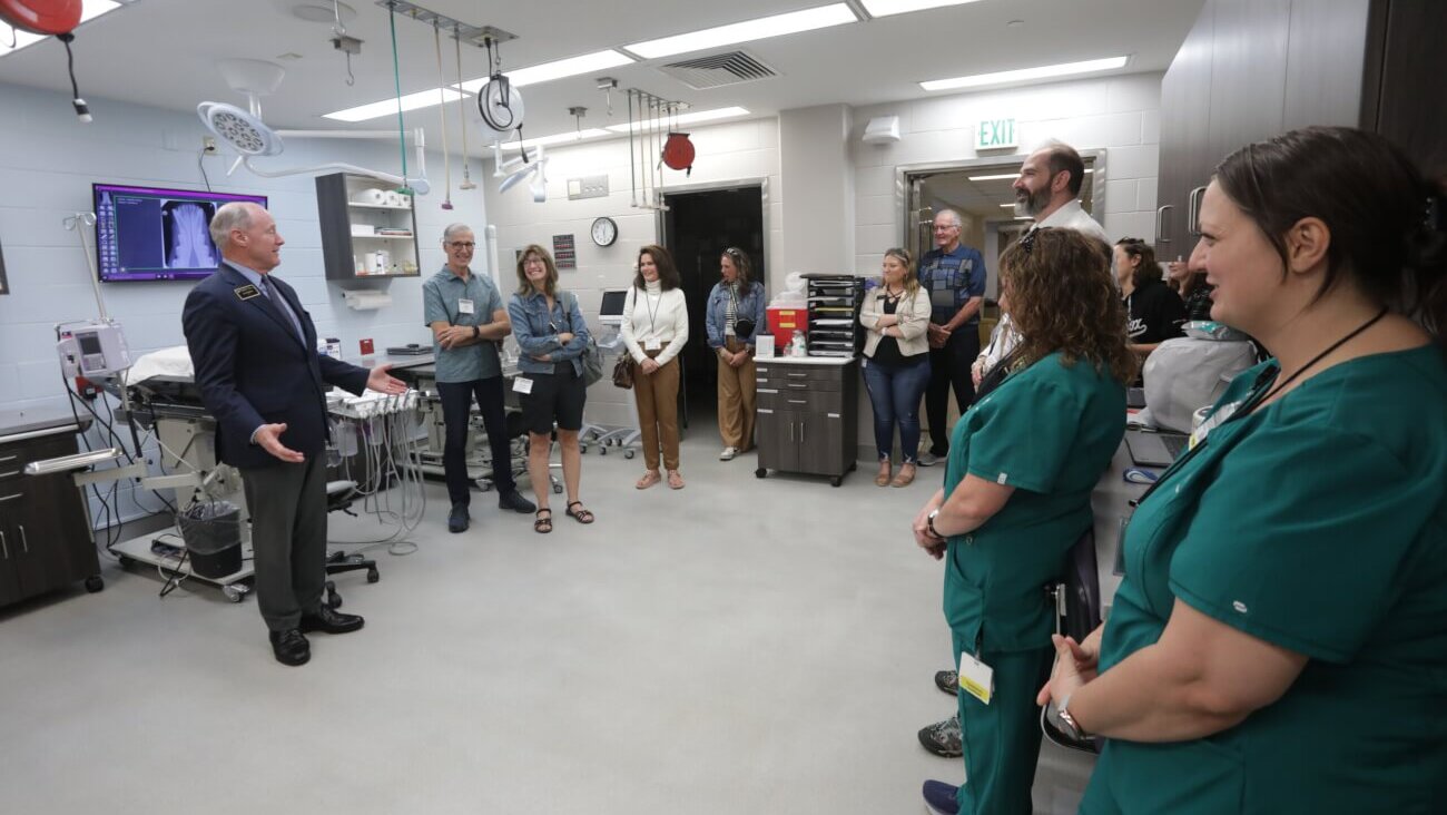 A large group of people are standing on the right side of the photo, observing a man positioned on the left side of the photo speak to them. The man is wearing a black suit with his arms out.