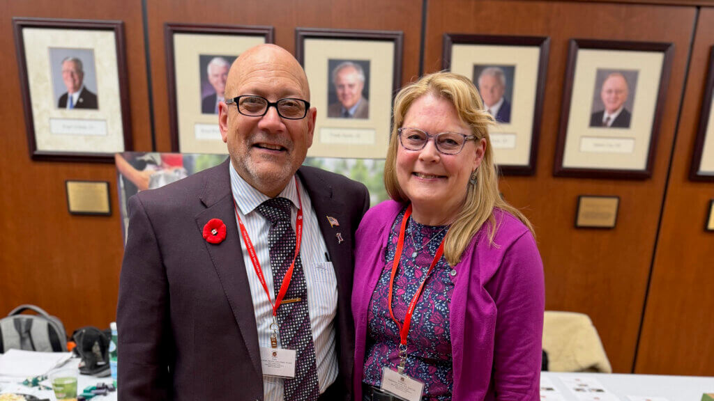 two people standing in front of a wall featuring awards. The man on the left is wearing a jacket with a red floral button. The woman on the right is wearing a purple shirt and jacket and has long, blonde hair.