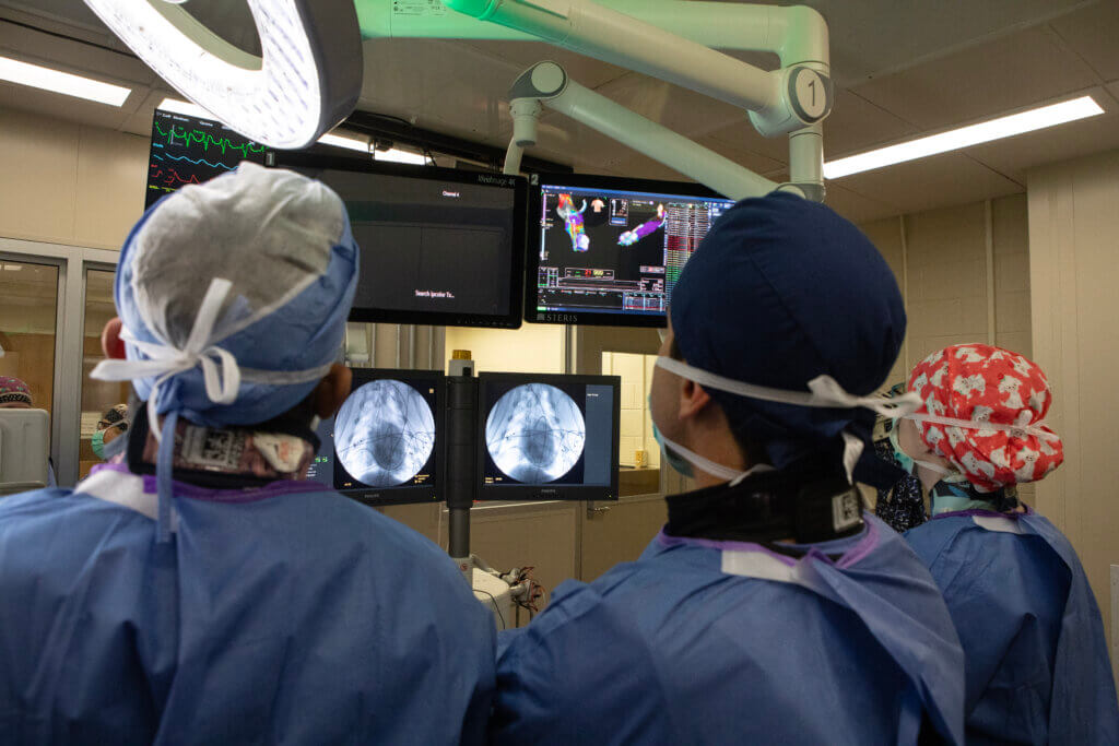 Two medical professionals in scrubs are observing monitors showing electrocardiogram readings and cardiac endoscopy images in an operation room, with their backs turned to the camera.