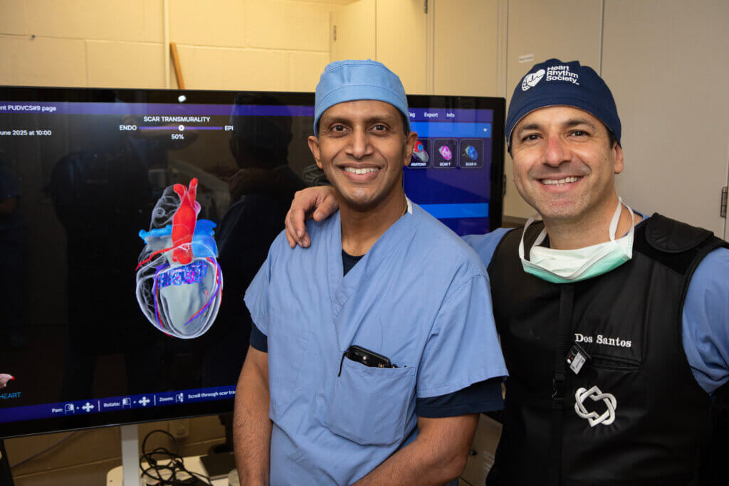 Two men in medical scrubs are facing the camera smiling. Behind them is a monitor showing a diagram of a heart.