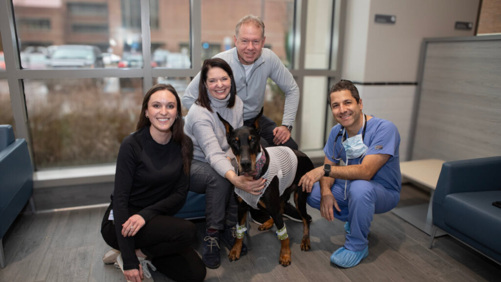 A group of people, including a veterinarian in scrubs, are posing in front of a window. In the middle of the group of people, there is a large black dog.
