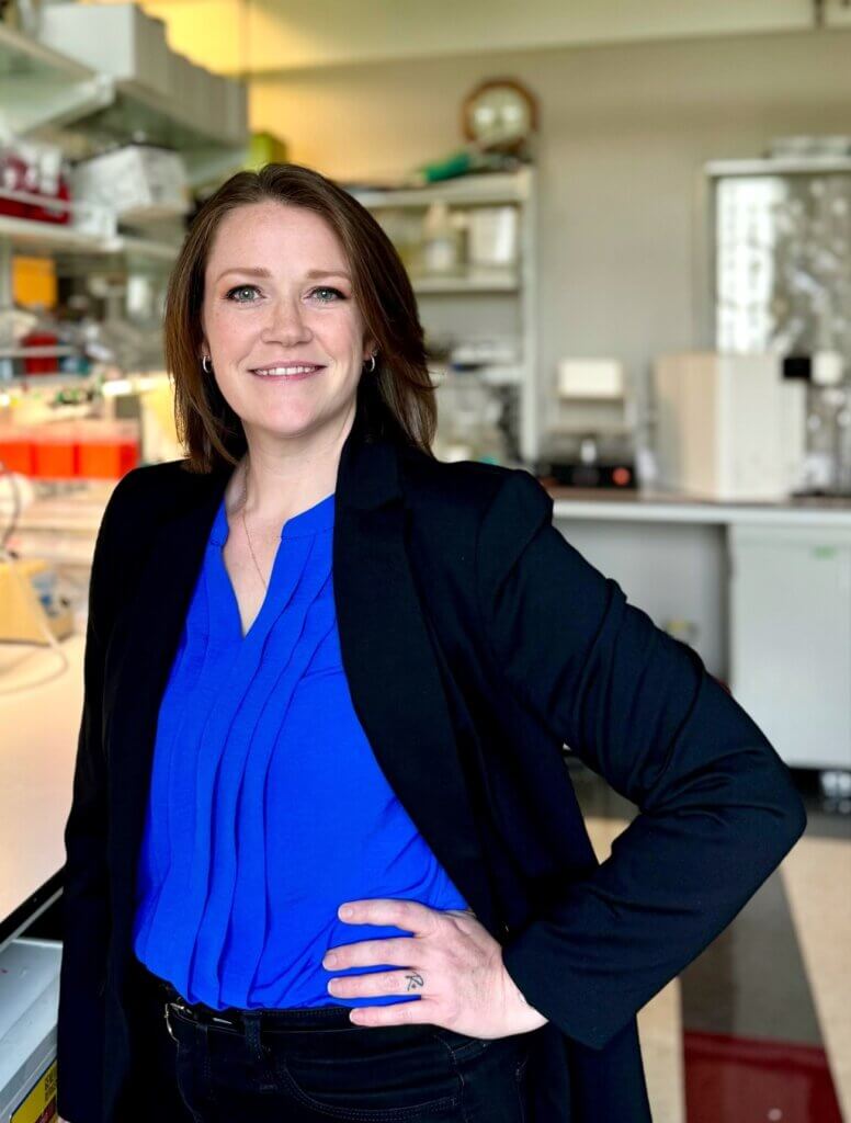 portrait of a woman in a blue shirt and black jacket, the background is a research lab setting