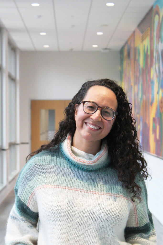 portrait of a woman wearing a blue and white sweater and glasses, standing in a hallway with natural lighting