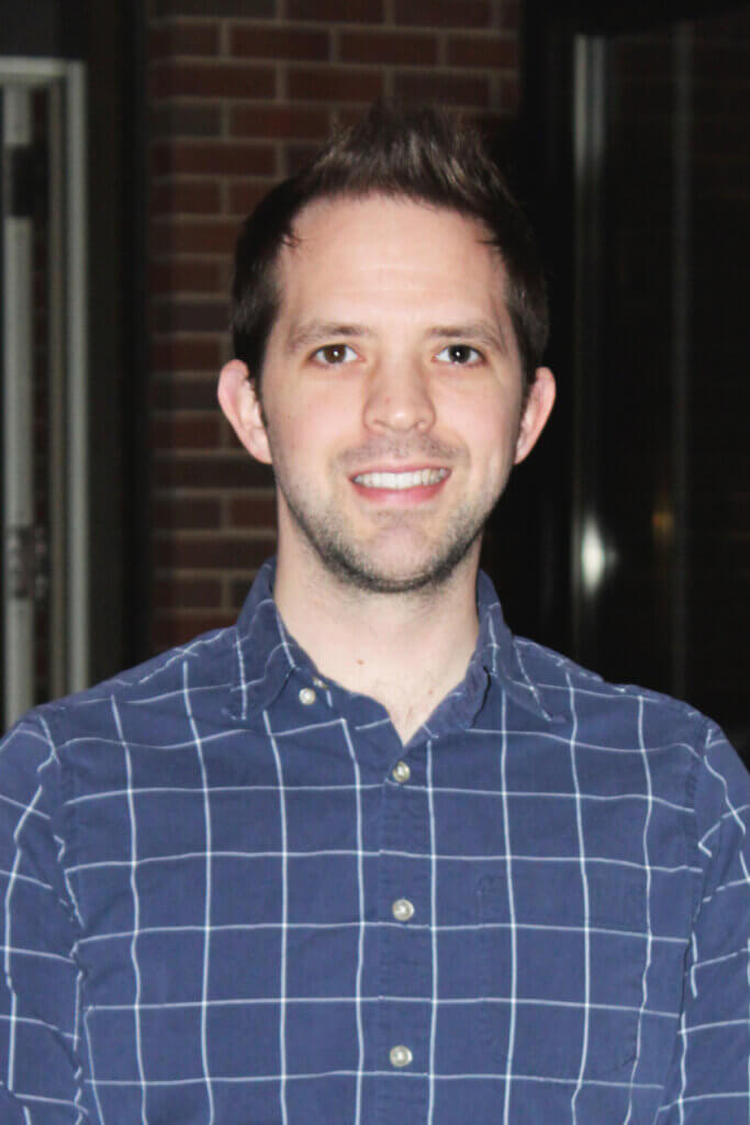 a portrait of a man wearing a blue and white plaid shirt in front of a red brick background