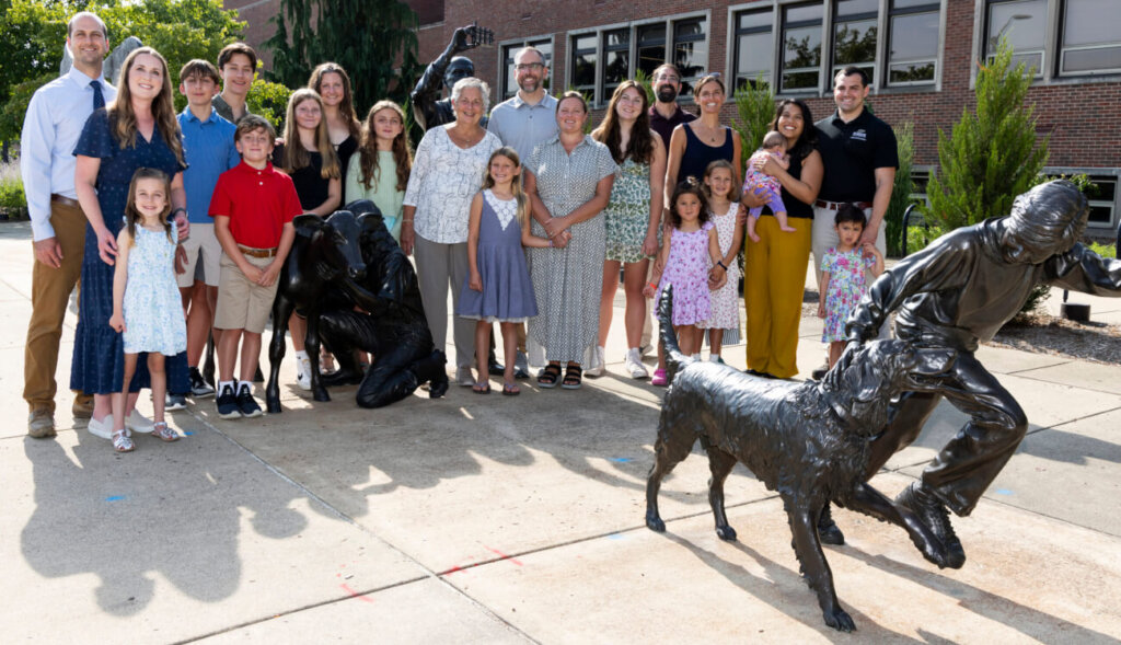 A large group of people, including all ages, is standing on pavement outdoors. There is a sculpture of a boy running next to a dog in front of the group of people.