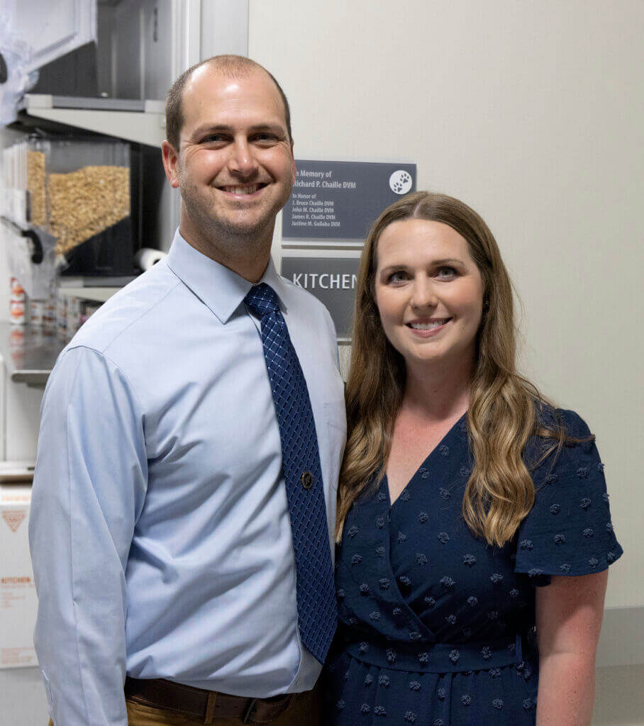 A man on the left and woman on the right are posed together. The man is wearing a blue dress shirt and the woman is wearing a dark blue dress