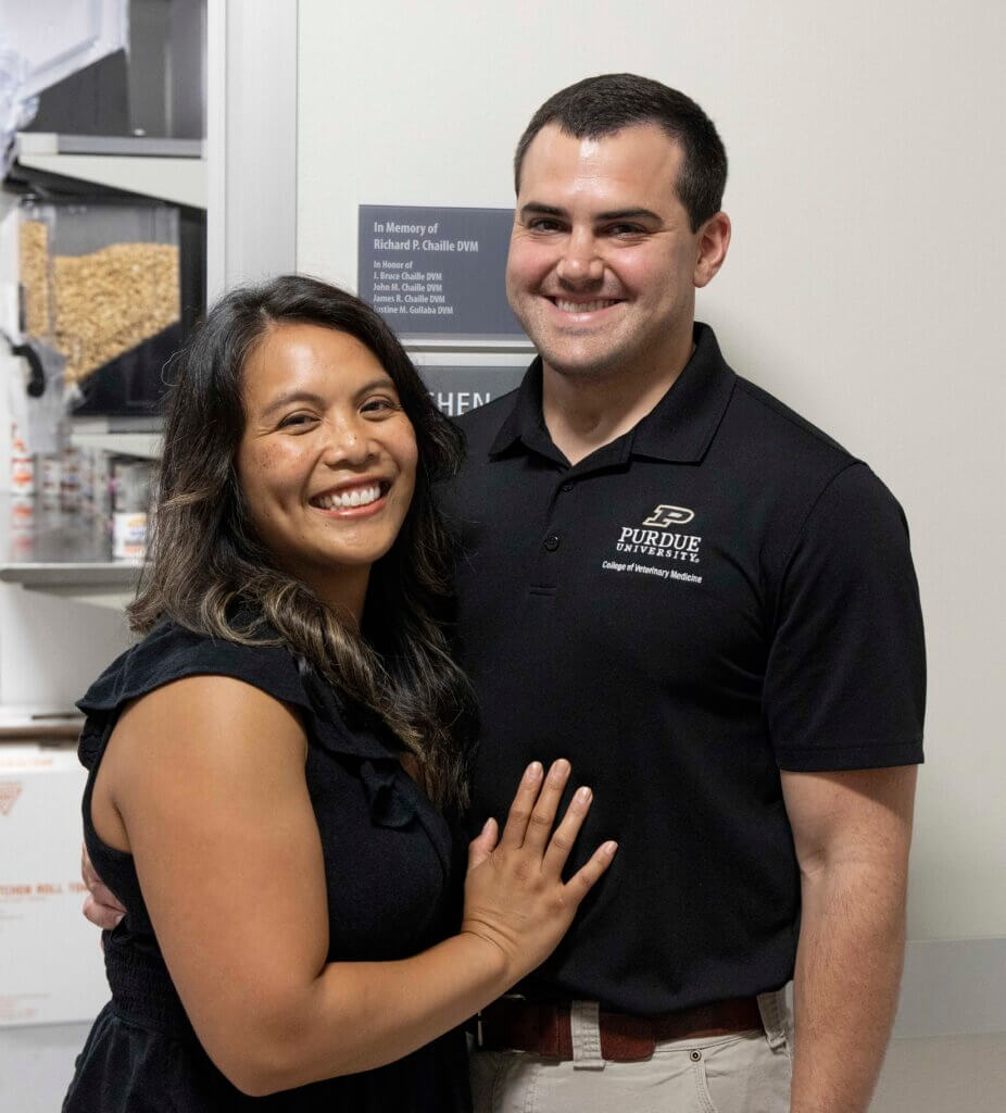 A woman and man are posed together with the woman resting her hand on the man's torso. Both people are wearing black