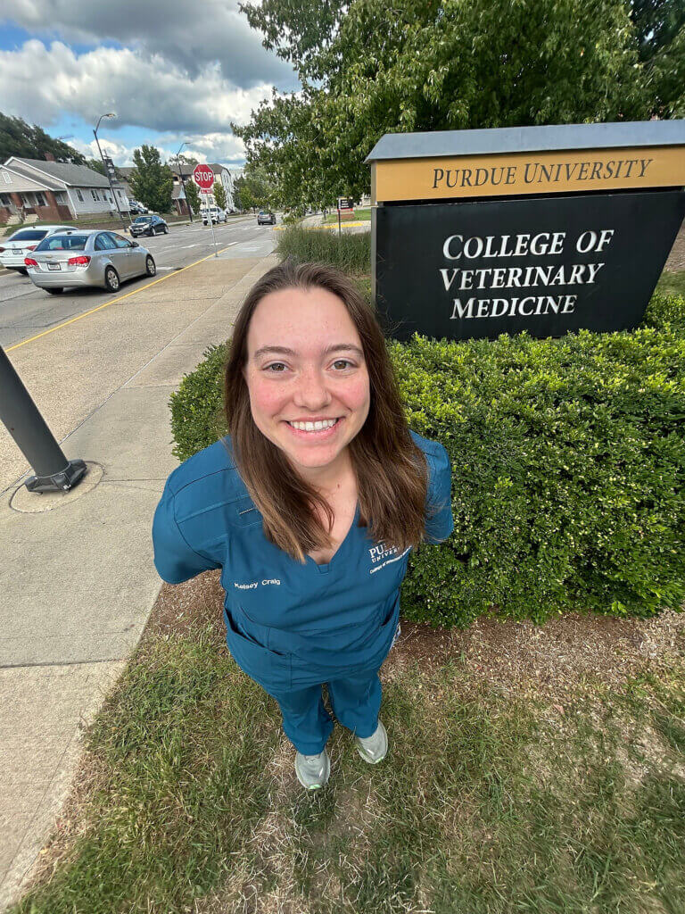 outdoors, a female student in dark blue scrubs smiling at the camera, with a Purdue University College of Veterinary Medicine sign behind her
