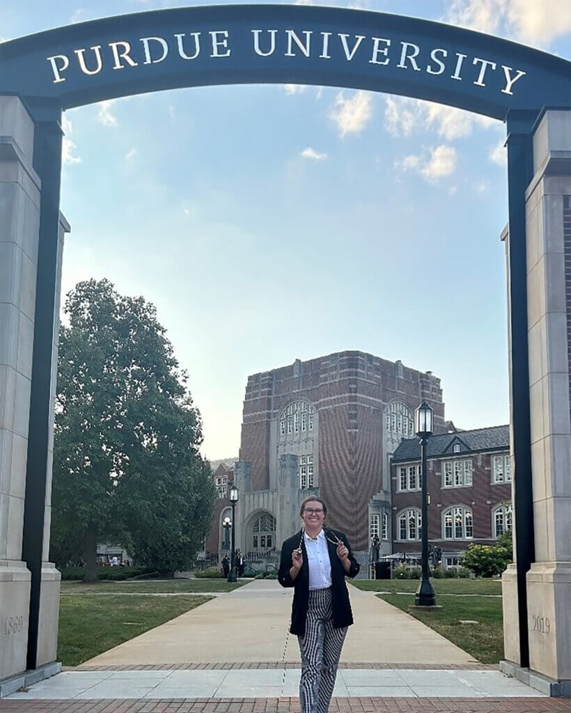 A female dressed in a black jacket and striped pants stands smiling under an arch that says "PURDUE UNIVERSITY" with a large brick building in the background