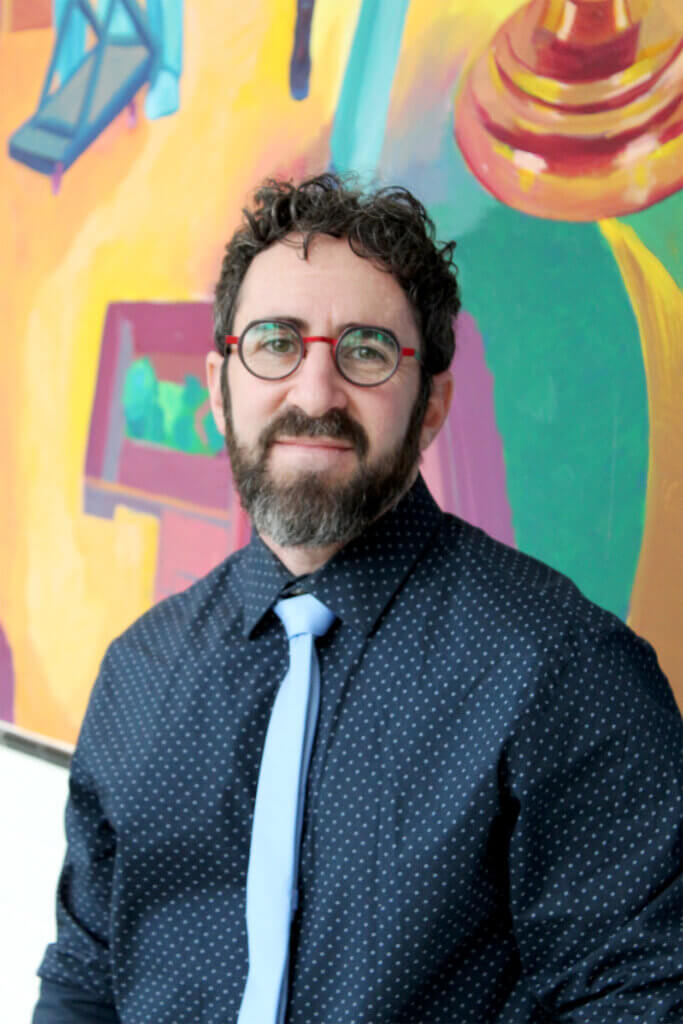 portrait of a man wearing a dark blue shirt with a light blue tie and glasses, standing in front of a colorful wall