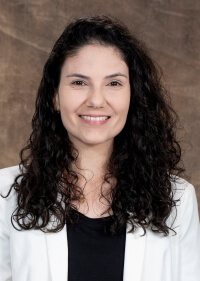 portrait of a woman with long dark hair, wearing a black shirt and a white jacket, posed in front of a brown background