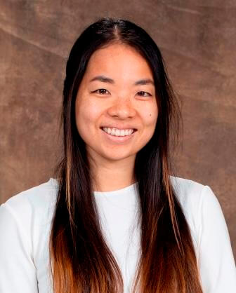 portrait of a woman who has long brown hair, wearing a white shirt and is smiling in front of a brown background