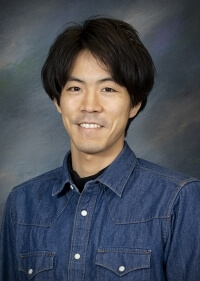 portrait of a man with dark hair wearing a blue button up shirt in front of a dark gray background