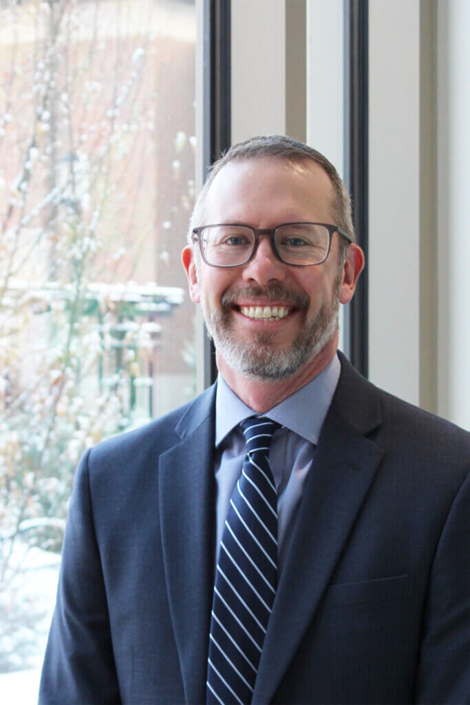 portrait of a man smiling, facing the camera in front of a snowy background. The man is wearing a dark suit jacket and a blue striped tie