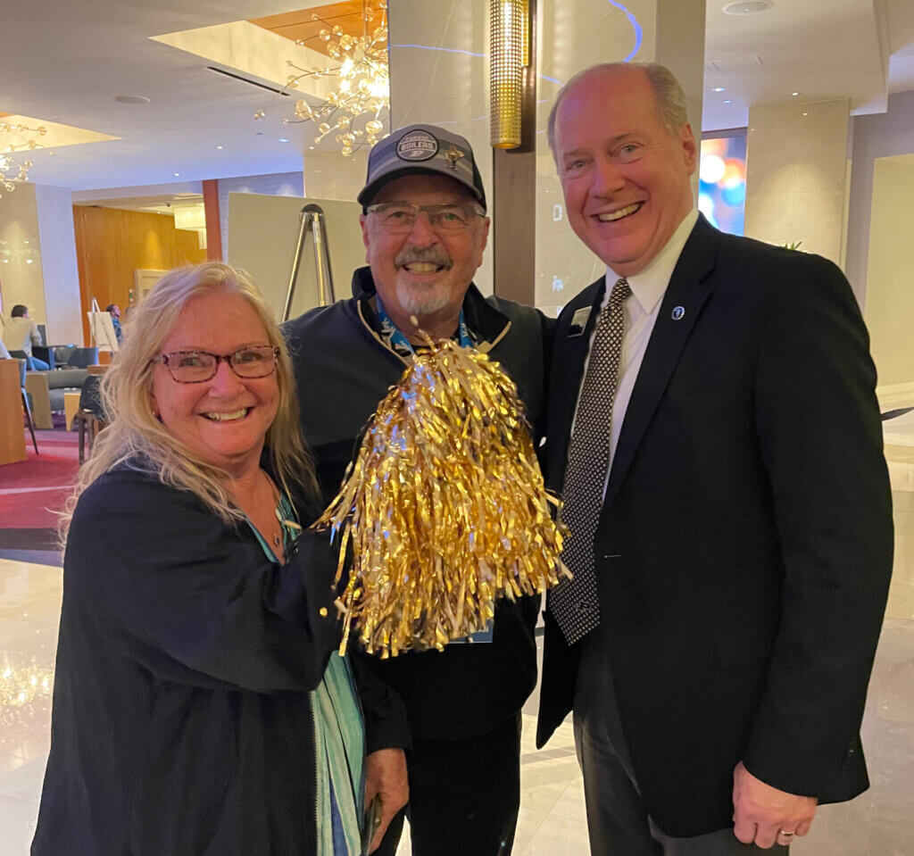 Three smiling adults pose together in a hotel lobby, with the woman on the left holding a gold pom-pom; one man wears a Purdue cap and lanyard, and the other is dressed in a suit and tie.