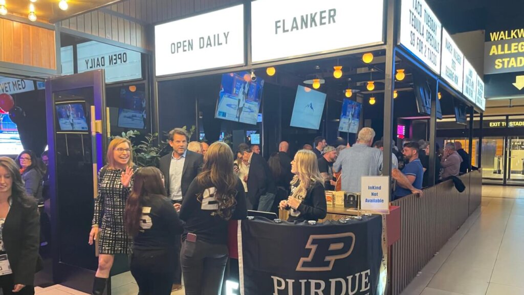 Guests gather around a Purdue University–branded table outside a bar area with illuminated “Open Daily” and “Flanker” signs, while people chat and watch sports on overhead TVs in a busy indoor venue.