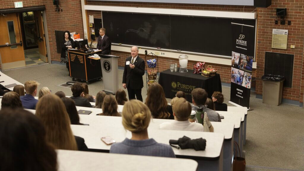 A man in a black suit is speaking to students in a tiered floor lecture hall.