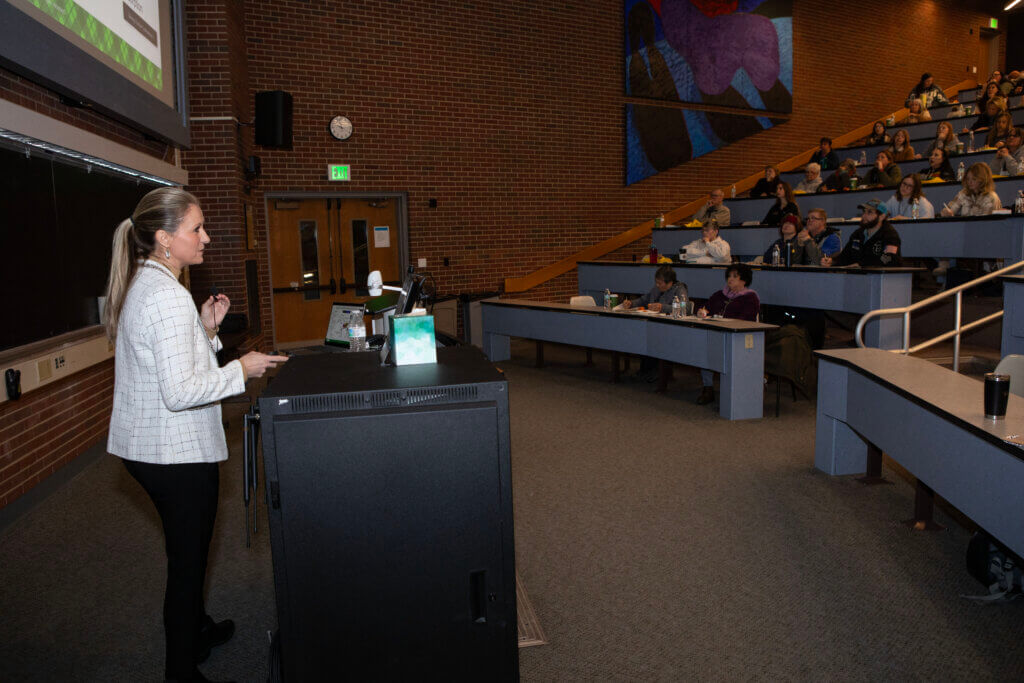 A woman wearing a white jacket is standing at the front of a lecture hall, giving a presentation to a large crowd