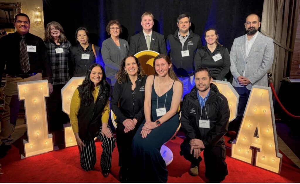 A group of people wearing business casual clothing is gathered around decorative letters for a photo. Behind them is a large black curtain.
