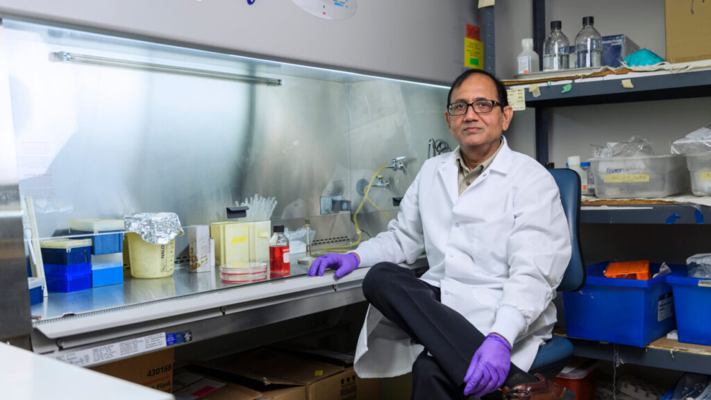 man in a laboratory setting, wearing a white coat and gloves and sitting in front of a counter with various scientific and laboratory items