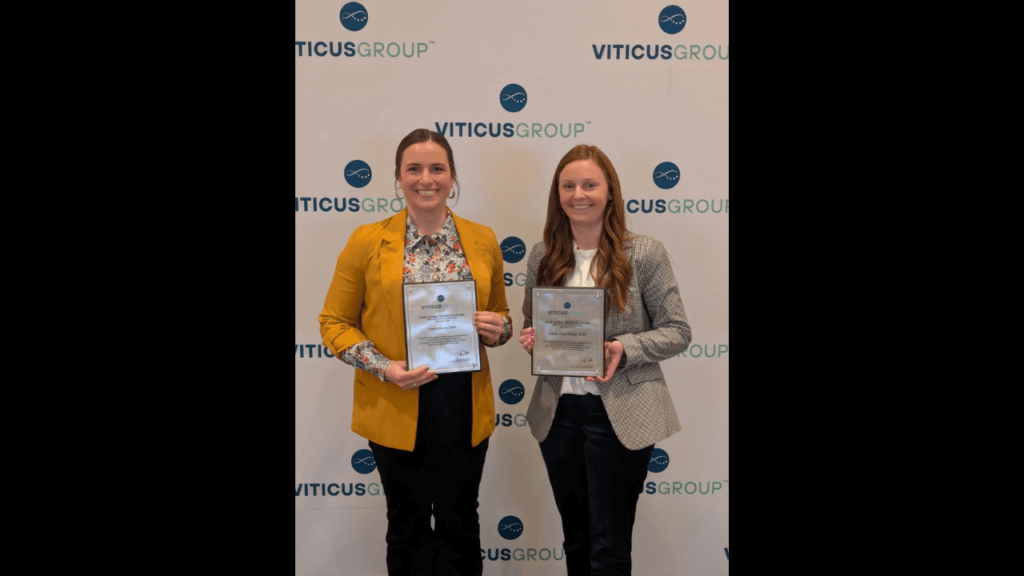 Two women are standing and holding awards. The woman on the left is wearing a yellow blazer and the woman on the right is wearing a gray blazer