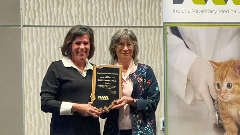 Two women are standing holding a plaque shaped as the state of Indiana. The background includes a banner with an orange cat.