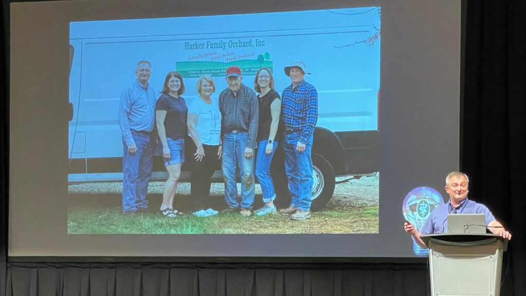 A man is standing at a podium in front of a projector screen that displays a slide of six people.