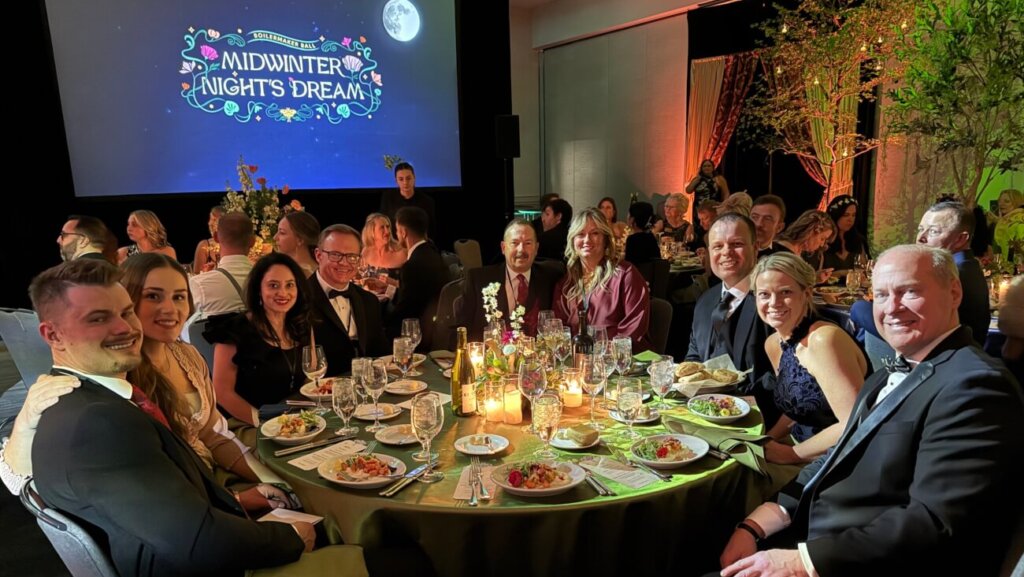 A group of people are wearing black-tie attire and sitting around a dinner table. There is a projector behind them displaying a winter graphic.