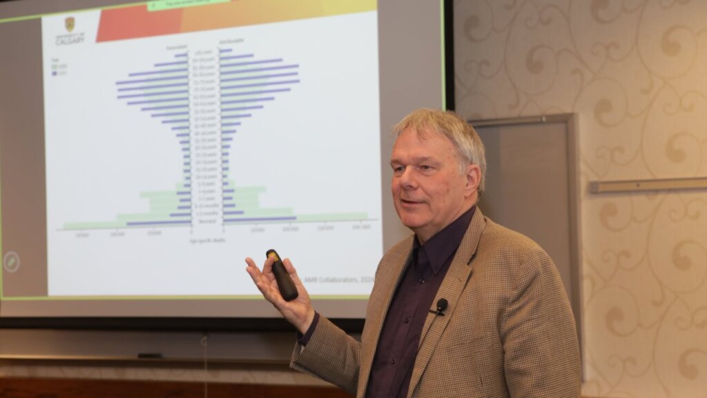 A man wearing a tan suit jacket appears to be giving a presentation in front of a projector screen