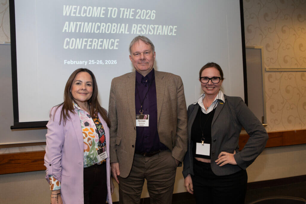 Three individuals are dressed in business attire and are standing in front of a projector screen that reads "Welcome to the 2026 Antimicrobial Resistance Conference"