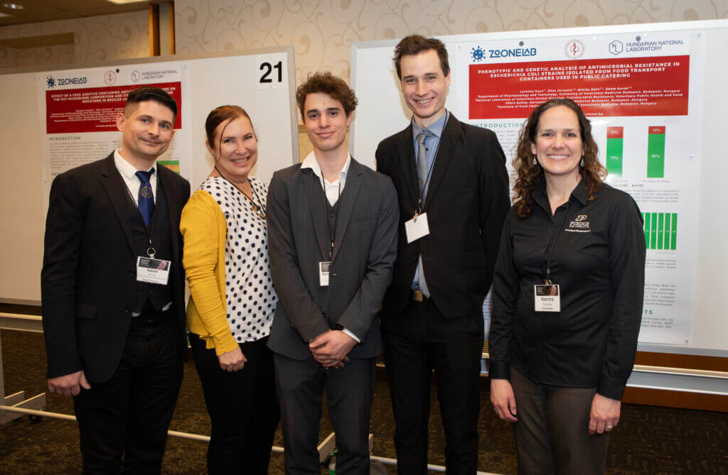 Five people are standing in a line, all dressed in business attire and positioned in front of research posters.