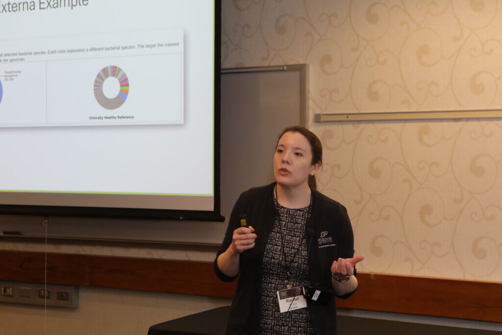 A woman wearing a black shirt appears to be giving a presentation in front of a projector screen