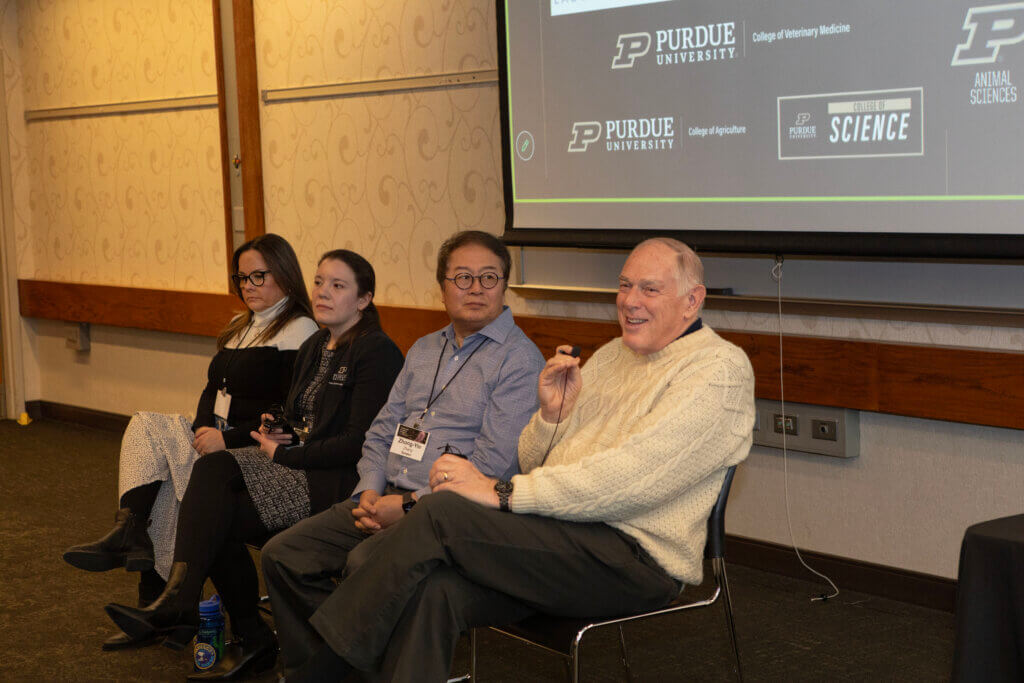 Four individuals are seated in front of a projector screen. One individual is holding a microphone and is wearing a yellow sweater.